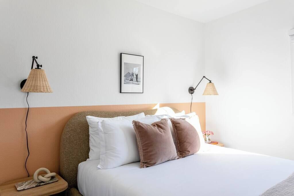A neatly made bed with white and brown pillows, a framed picture on the wall, and two wall-mounted lamps.