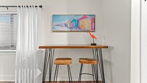 Een kamer met een wit gordijn, een schilderij van kleurrijke gebouwen, een houten tafel met twee krukken, en een raam met jaloezieën.