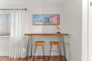 A room with a white curtain, a painting of colorful buildings, a wooden table with two stools, and a window with blinds.