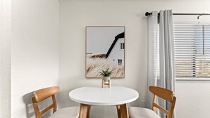 Ein Essbereich mit einem runden weißen Tisch, zwei Holzstühlen mit gepolsterten Sitzen, einem gerahmten Bild an der Wand und einem Fenster mit Jalousien.