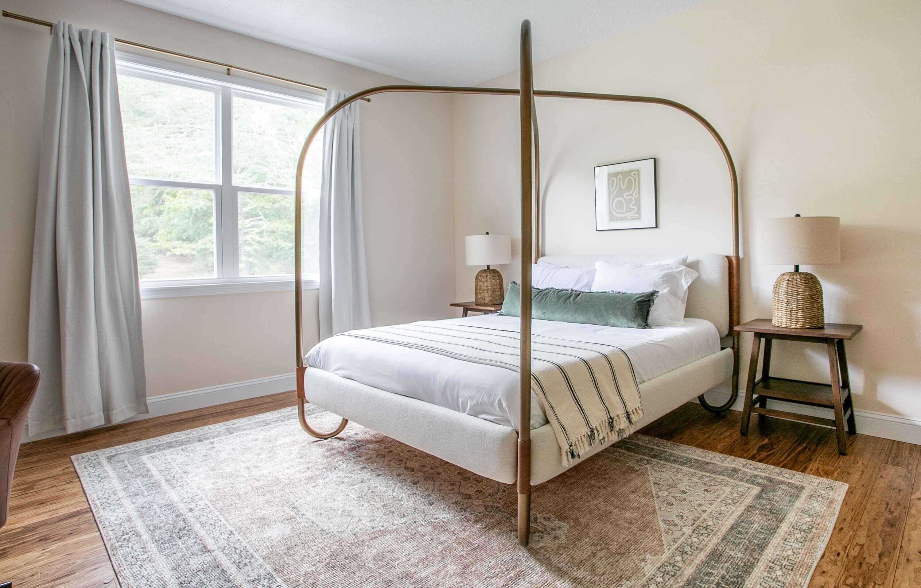A four-poster bed with a white and green color scheme, a small bedside table with a lamp, and a large window with curtains.
