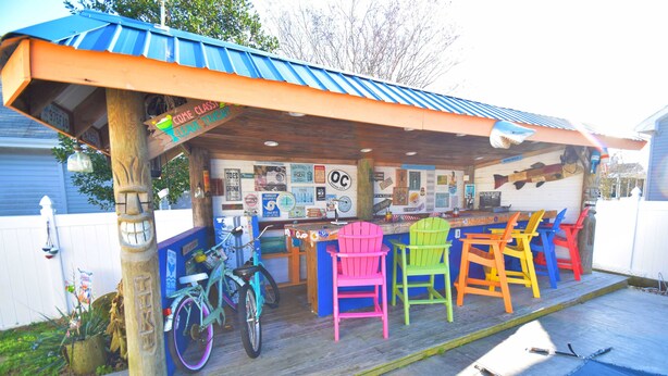 A covered outdoor seating area with colorful Adirondack chairs, a bicycle, and a wooden deck.