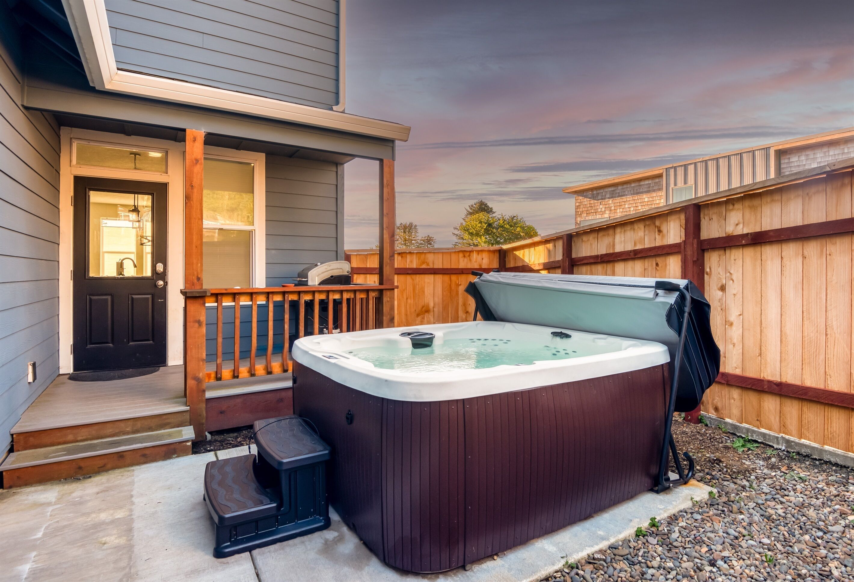 Un bain à remous installé dans un jardin, avec une terrasse en bois, une porte et une petite marche menant à l’entrée.