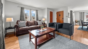 A living room with a brown leather sofa, a dark grey sofa, a wooden coffee table, and a grey rug. There are two lamps on side tables, a potted plant on the coffee table, and a view of the outside through a window with curtains.