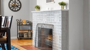 A fireplace with a brick pattern, a clock on the wall, and a small shelf with plants.