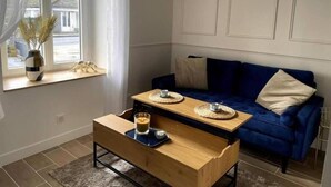 Una sala de estar con un sofá azul, una mesa de centro de madera, una mesita con una taza y un plato, un florero con plantas secas y un reloj de pared.