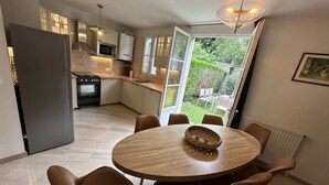 A modern kitchen with a dining area, featuring a round table with chairs, a built-in oven, and a view of a garden through a glass door. - Appartement Avec Jardin en Plein Cœur de Barbizon (Barbizon)