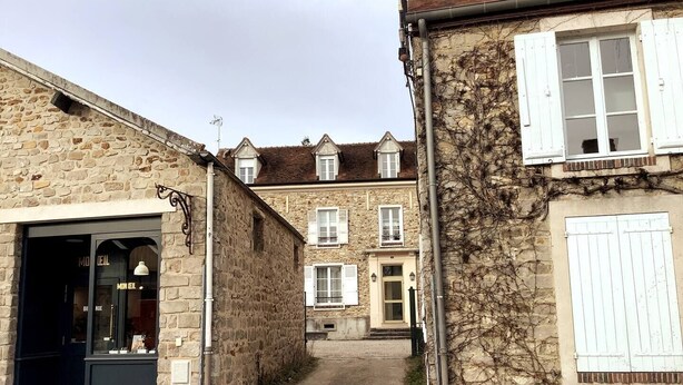 A narrow street with stone buildings, a shop with a glass door, and a building with white shutters. - Appartement Avec Jardin en Plein Cœur de Barbizon (Barbizon)