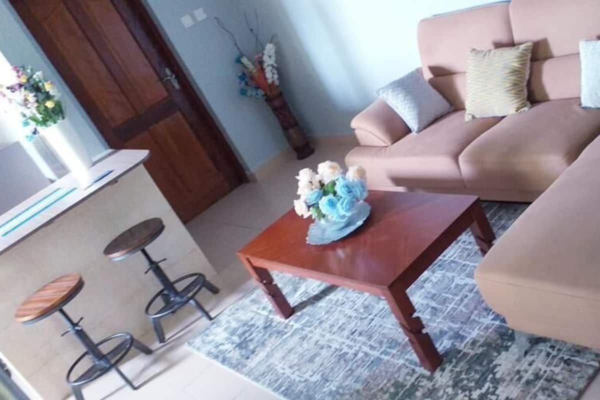 A living room with a beige sofa, a wooden coffee table with a floral arrangement, and a patterned rug. There are two bar stools and a small table with a vase of flowers.
