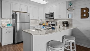 A modern kitchen with white cabinetry, stainless steel appliances, a marble countertop, and a breakfast bar with stools. - Dolphin Overlook - Beautiful oceanfront condo with pool (Carolina Beach)