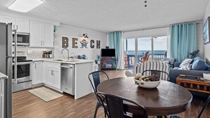 A modern kitchen with white cabinets, a dining area with a wooden table, and a view of the ocean through large windows. - Dolphin Overlook - Beautiful oceanfront condo with pool (Carolina Beach)