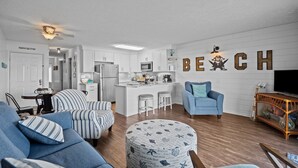 A modern living room with a sofa, ottoman, and a blue armchair. A kitchen with white cabinets and a bar area is visible. - Dolphin Overlook - Beautiful oceanfront condo with pool (Carolina Beach)