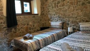 A room with two beds, each with patterned bedding and pillows, a stone wall, a window with a curtain, and a nightstand with a lamp. - Vivienda Vacacional Naguar Para 4 Personas en Triongo, Cangas de Onís (Triongo, Cangas de Onís)