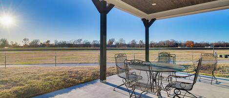 An outdoor patio with a table and chairs, a ceiling fan, and a view of a field and trees.