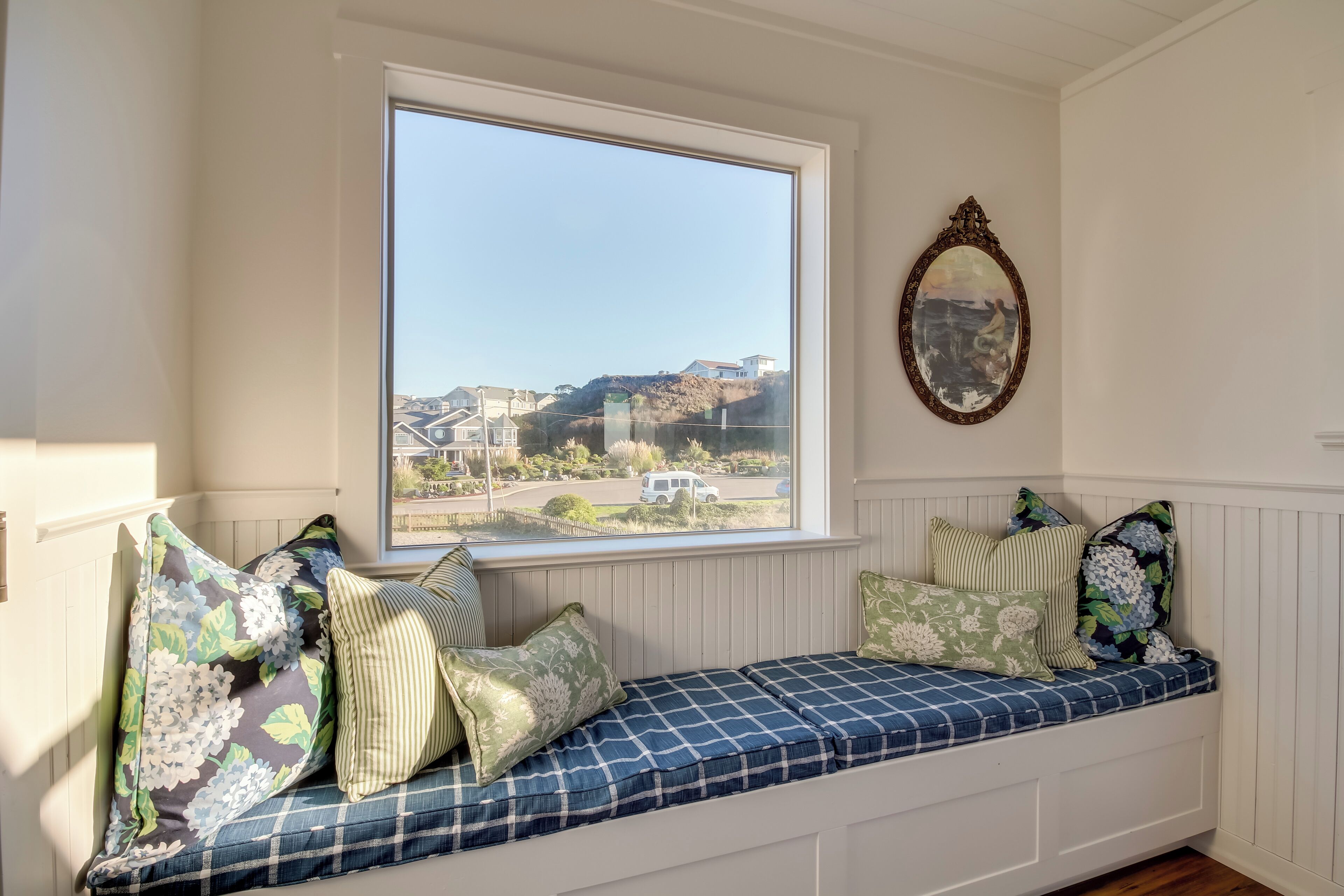 Ein Zimmer mit eingebauter Bank mit Kissen, einem großen Fenster mit Blick auf Häuser und eine Straße sowie einem Spiegel an der Wand.