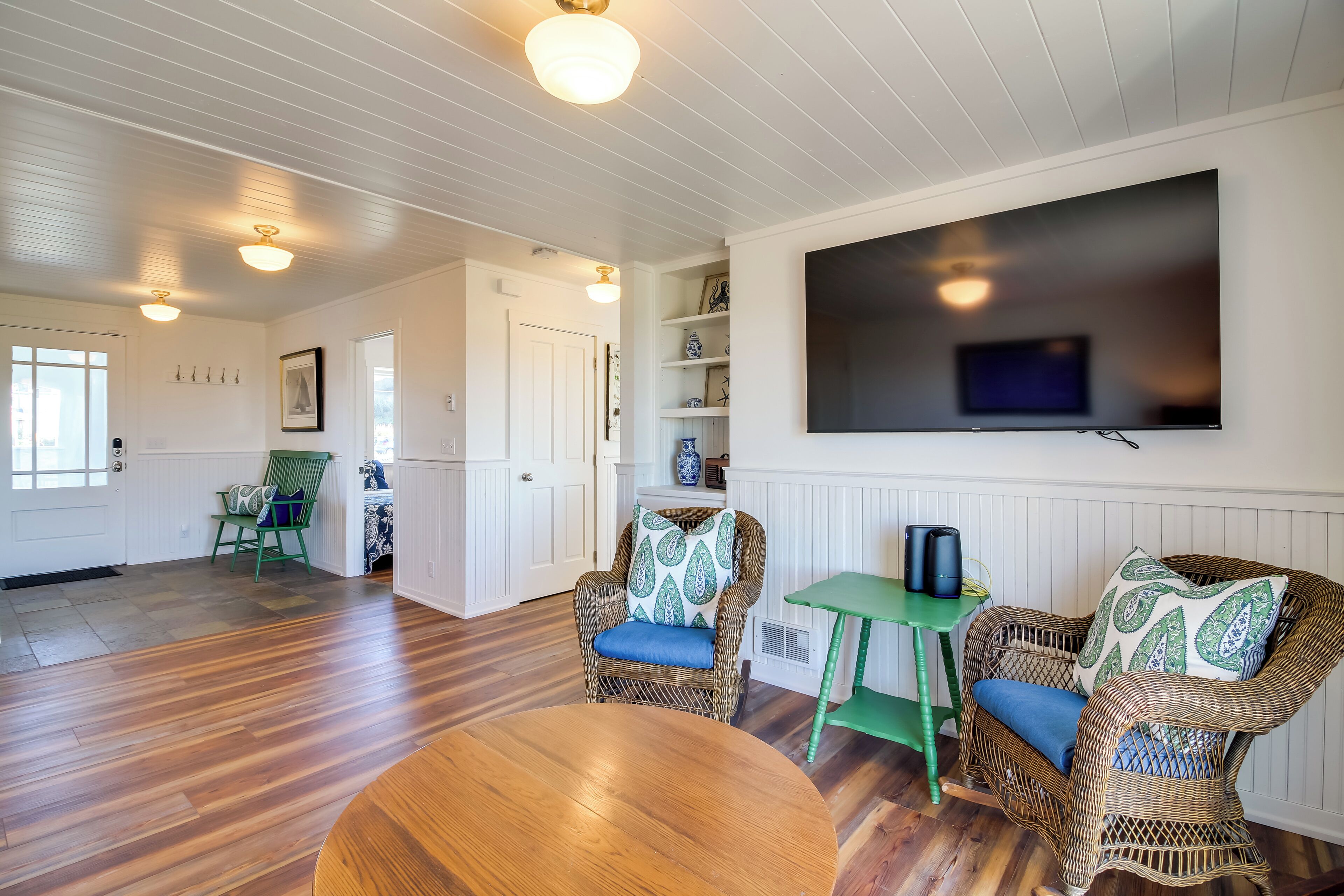 A spacious living room with a large flat-screen TV mounted on the wall, wicker furniture, and a wooden floor.