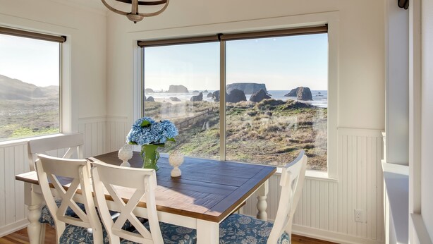 A dining area with a wooden table and white chairs, large windows with a view of the beach and rocks, and a chandelier. - Scenic Bandon Vacation Rental w/ Beach Access! (Bandon)