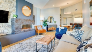 A modern living room with a stone fireplace, a flat-screen TV, and a comfortable seating area with a sofa and ottoman. - Downtown Winter Park Condo: Steps to Everything! (Winter Park)