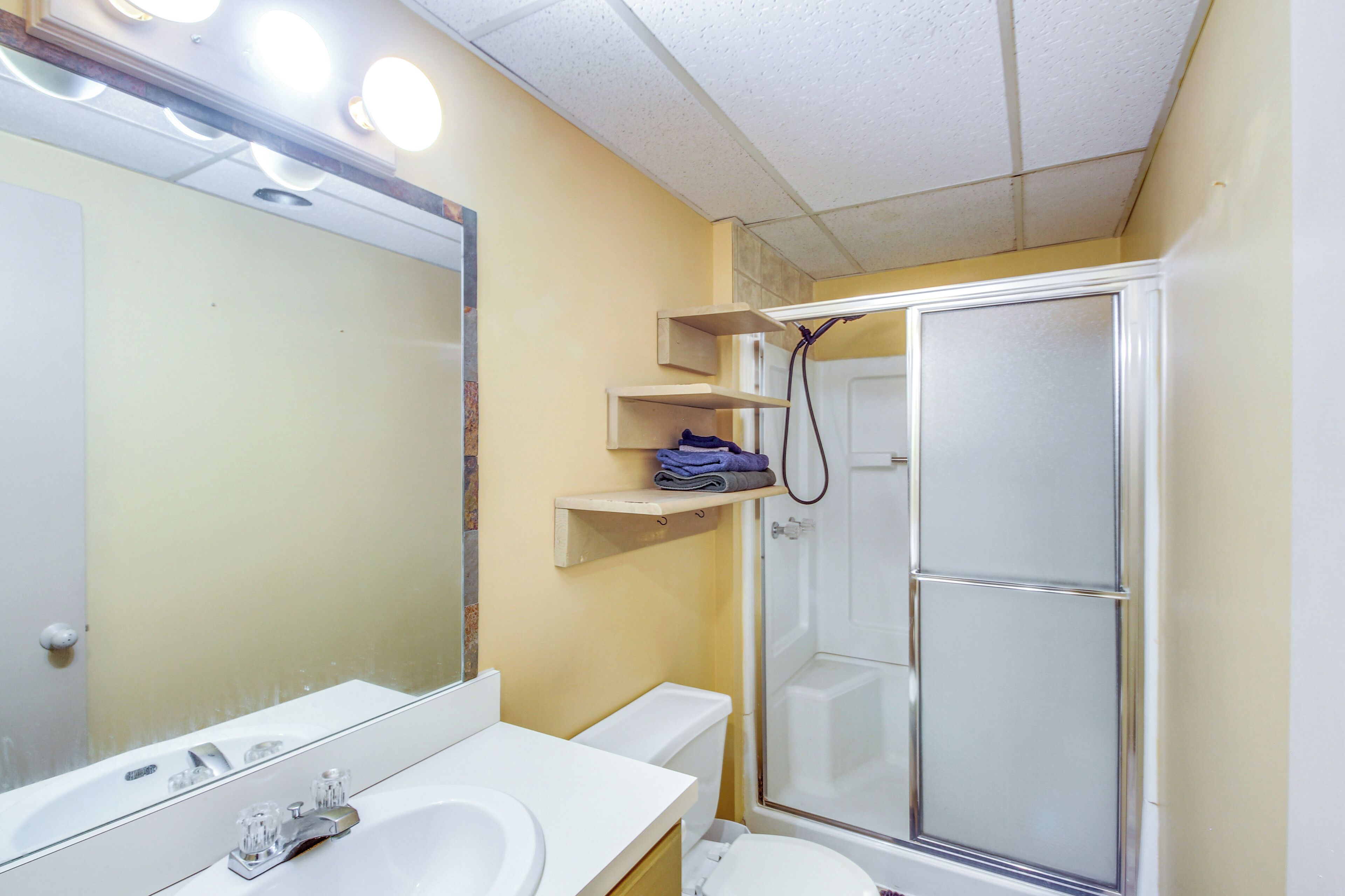 Un baño con lavamanos, espejo y retrete blancos. Hay repisas sobre el retrete y una cabina de ducha de vidrio.