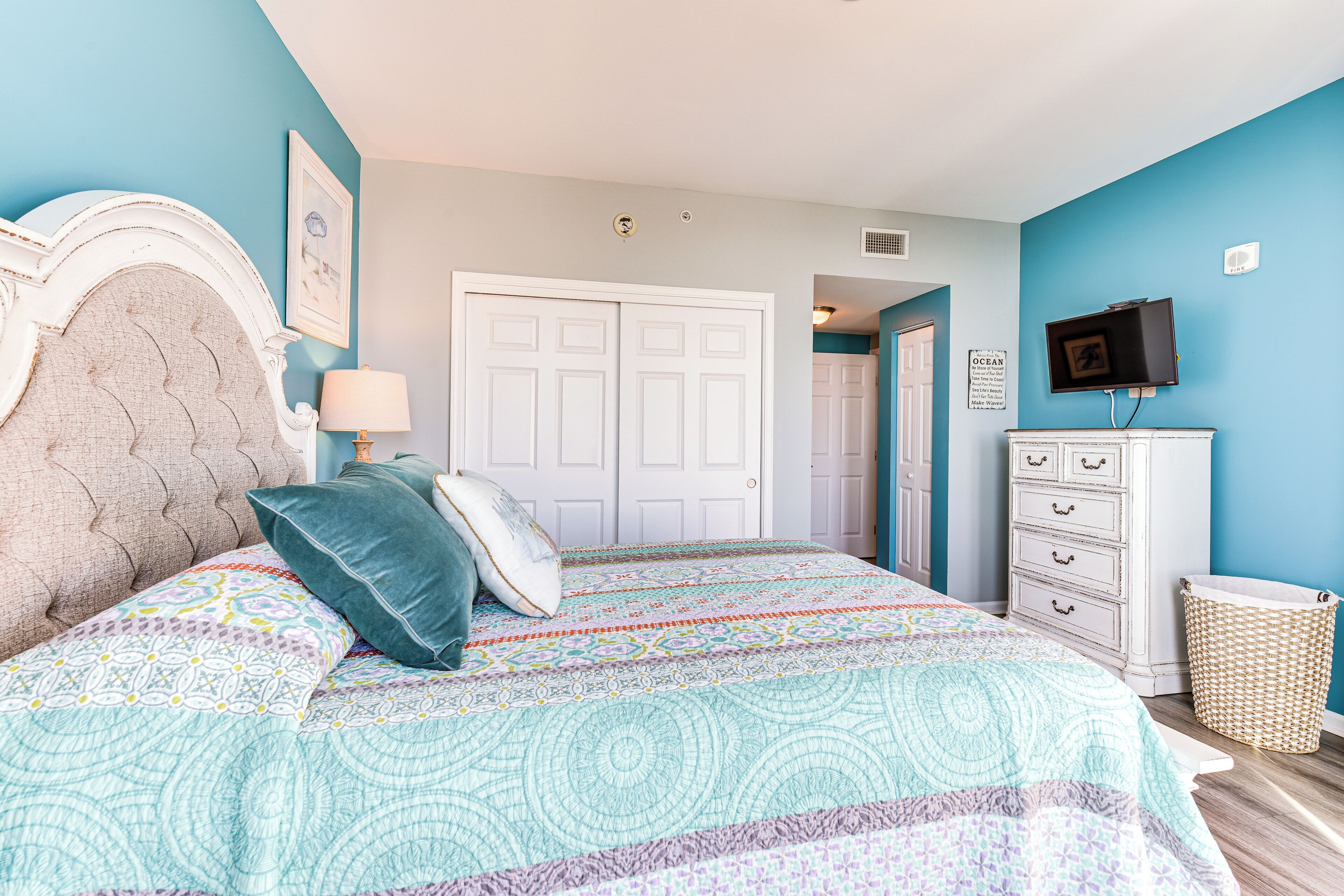 A bedroom with a large bed, a dresser, a television, and a door leading to another room.