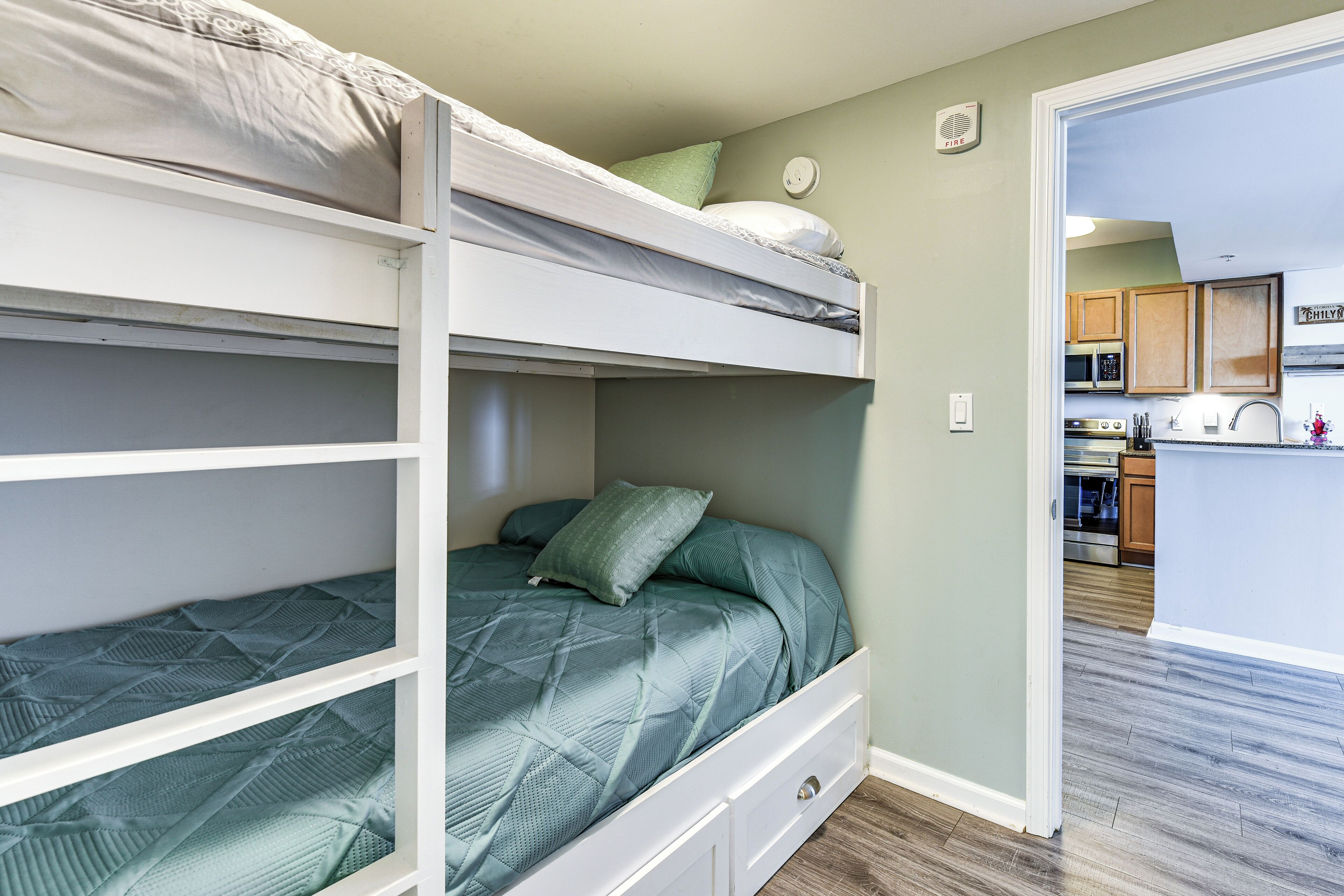 A bunk bed with a green bedspread and a pillow, a white dresser, and a kitchen area with wooden cabinets and a microwave.