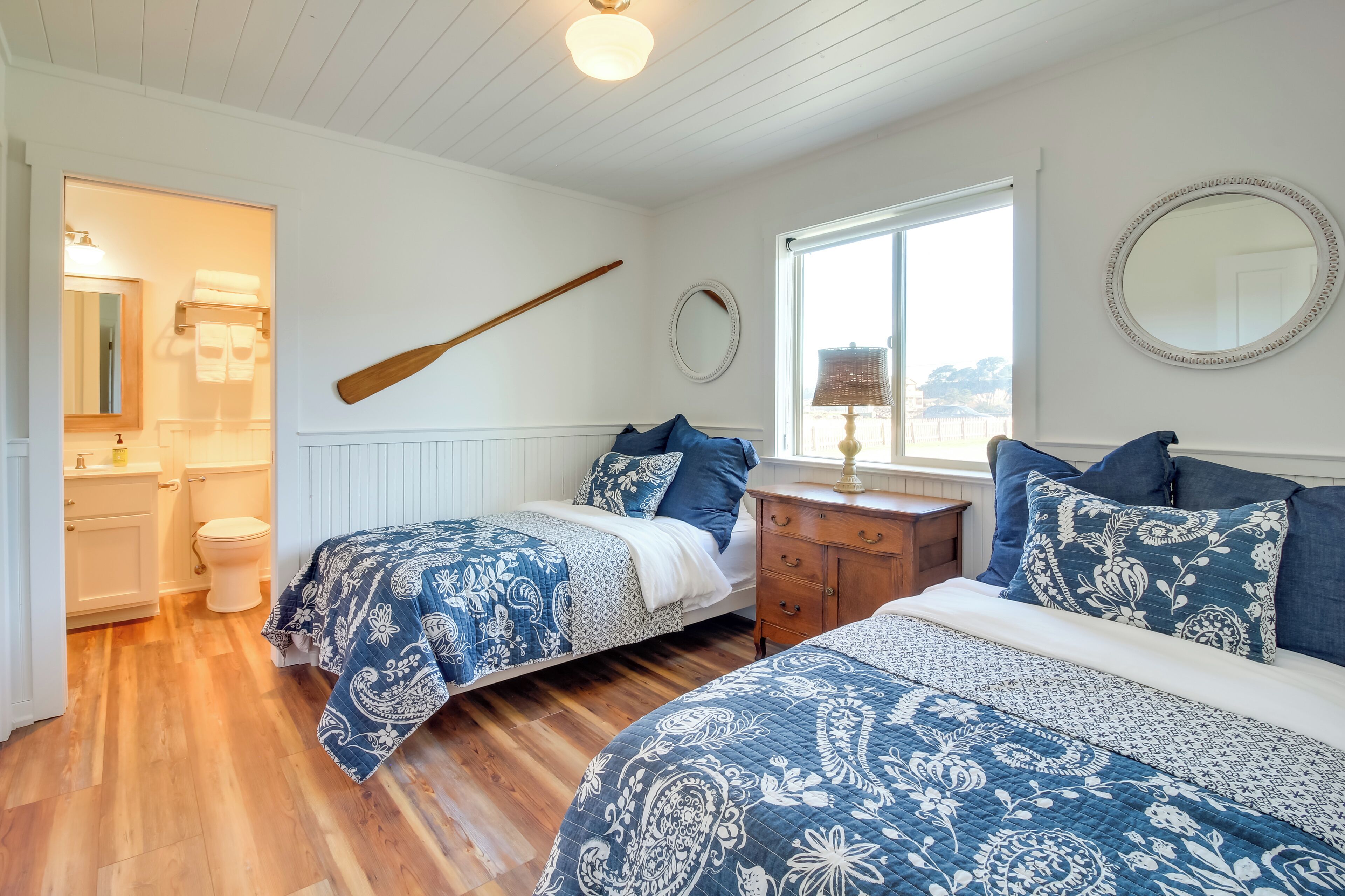 A bedroom with two beds, a wooden floor, a wooden nightstand, a mirror, and a window with a view.