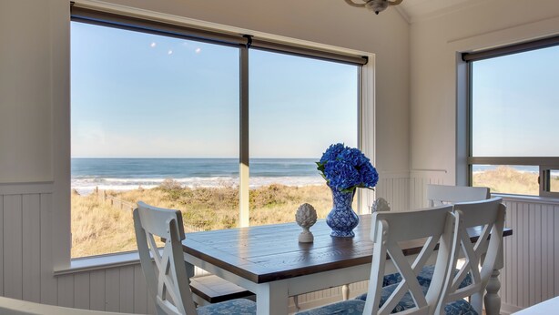 A room with a view of the ocean, a wooden table, white chairs, and a vase with blue flowers. - Beachfront Bandon Vacation Rental w/ Ocean View! (Bandon)