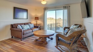 A living room with a brown leather sofa, a wicker chair, a wooden coffee table, and a large window with a view of a fence. - Beachfront Bandon Vacation Rental w/ Ocean View! (Bandon)