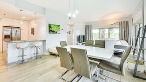 Une cuisine moderne dotée d’un réfrigérateur encastré, un coin repas avec une grande table et des chaises, et un espace détente avec un canapé et un téléviseur.