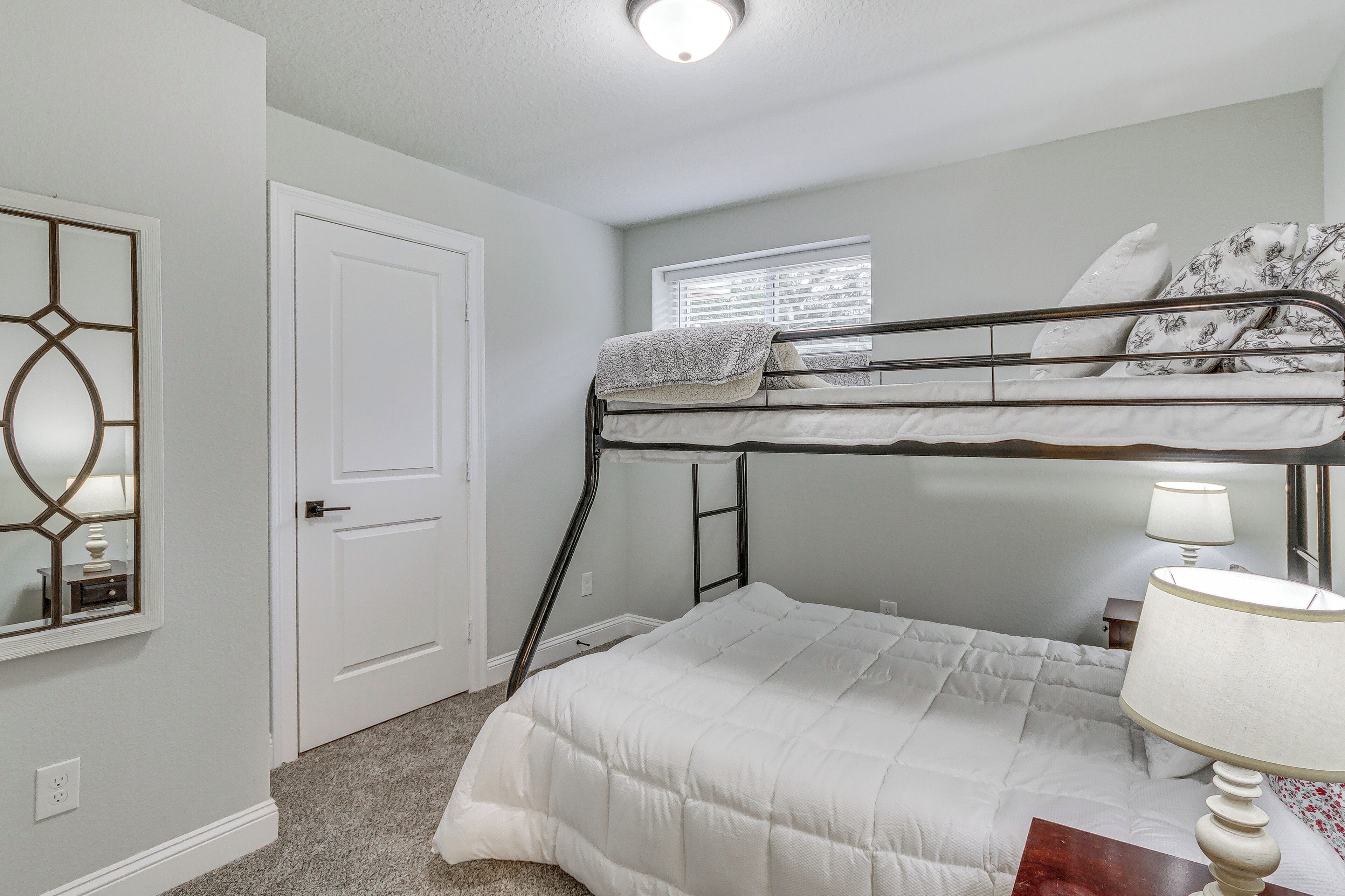 A bedroom with a bunk bed, a single bed, a bedside table with a lamp, a window with blinds, and a door.
