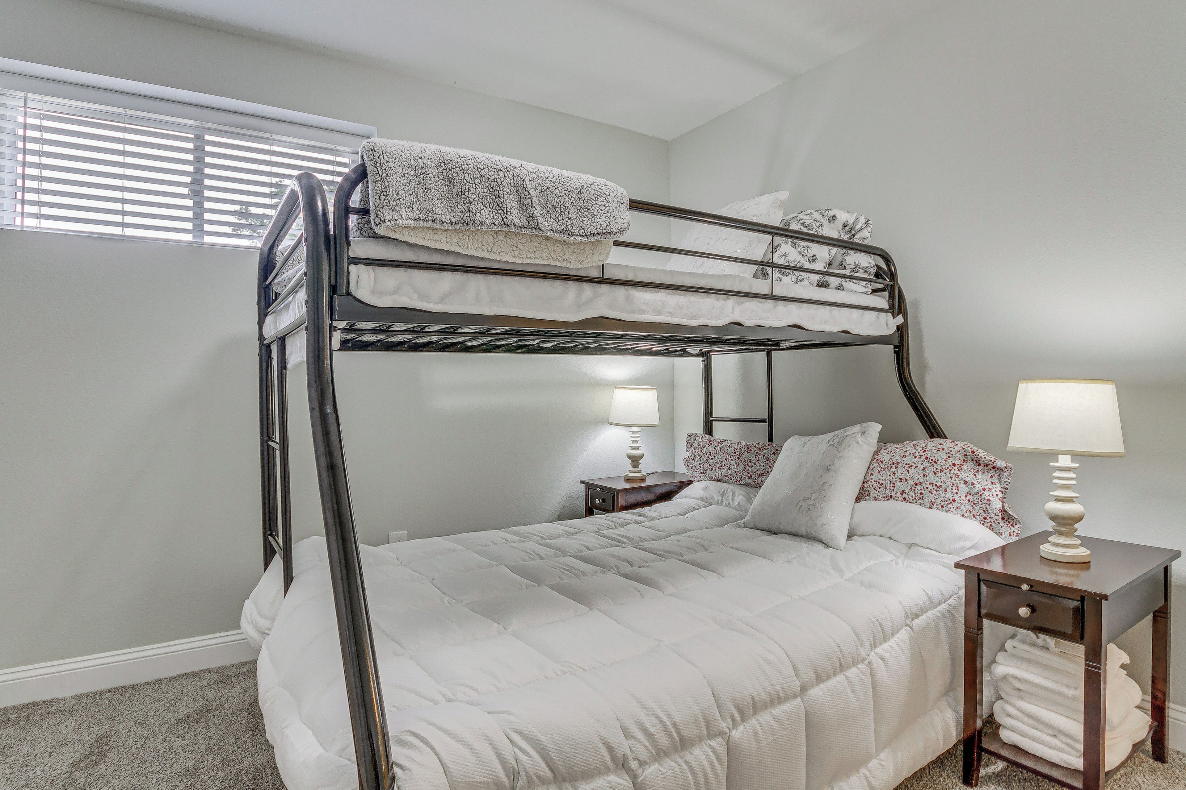 A bunk bed with a white comforter, a bedside table with a lamp, and a window with blinds.