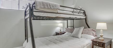 A bunk bed with a white comforter, a bedside table with a lamp, and a window with blinds.