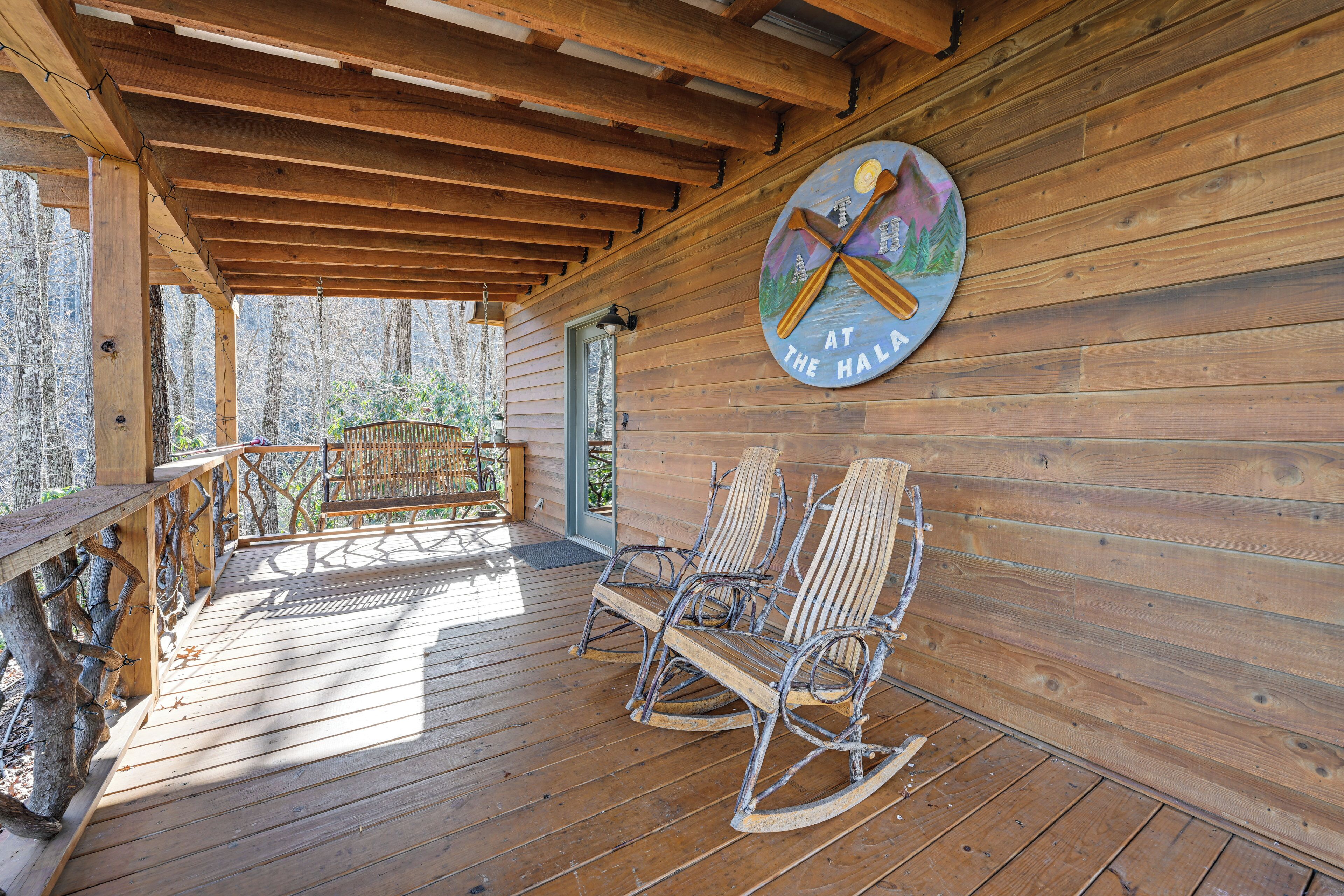 Un patio in legno con poltrone a dondolo e una panca, un muro di legno con un cartello circolare, e una vista sugli alberi.