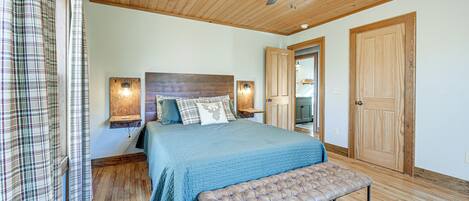 A bedroom with a wooden ceiling, a bed with a blue bedspread, a bench, and a door.