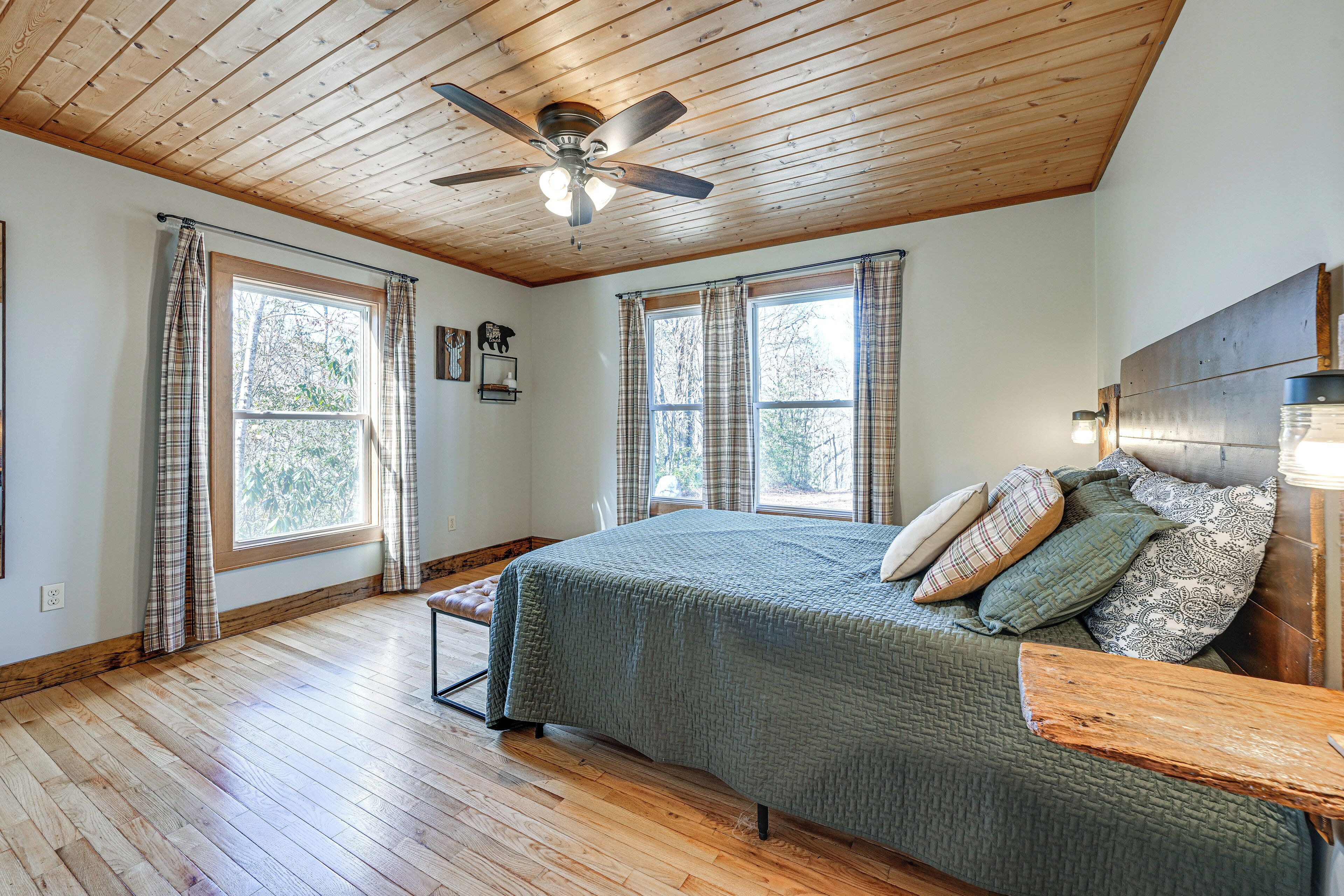 A bedroom with a wooden ceiling, a bed with multiple pillows, and two windows with curtains.