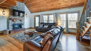 A spacious living room with a stone fireplace, leather sofas, a wooden staircase, and large windows.