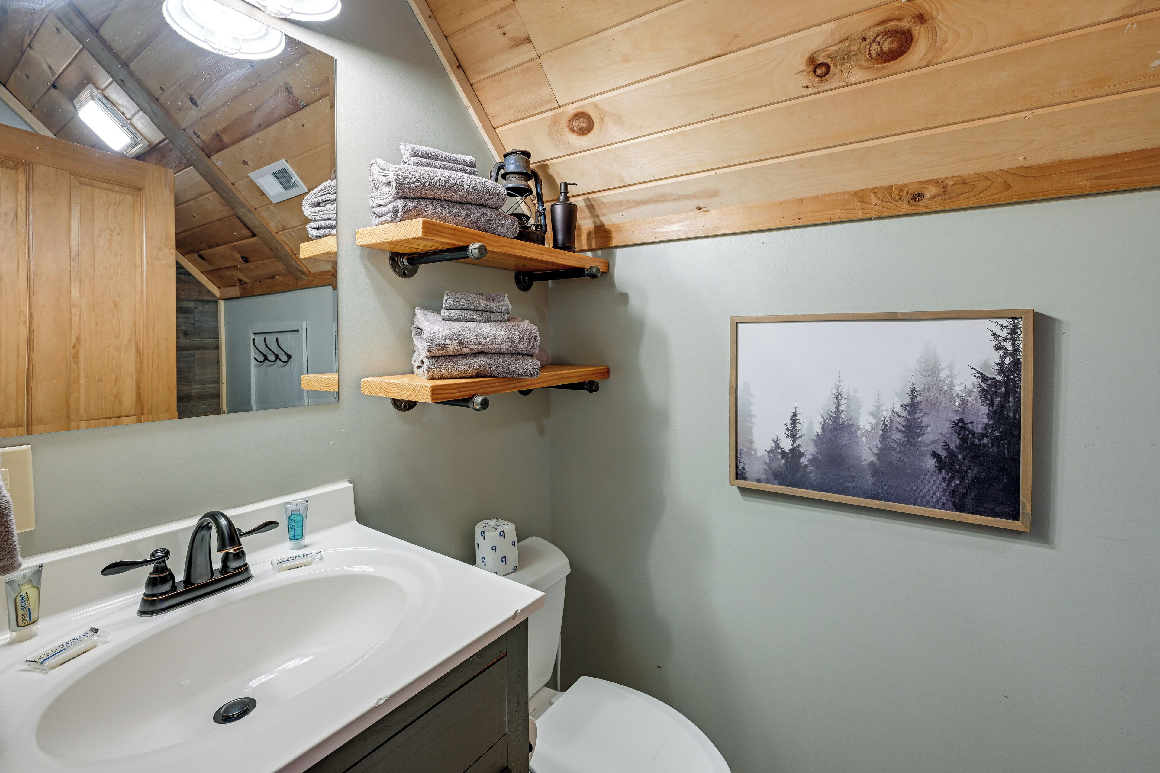 A bathroom with a white sink, wooden shelves above the toilet holding towels, a framed picture of trees, and a wooden ceiling.