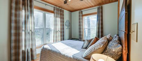 A bedroom with a large bed, plaid curtains, a ceiling fan, and a window with a view of trees.