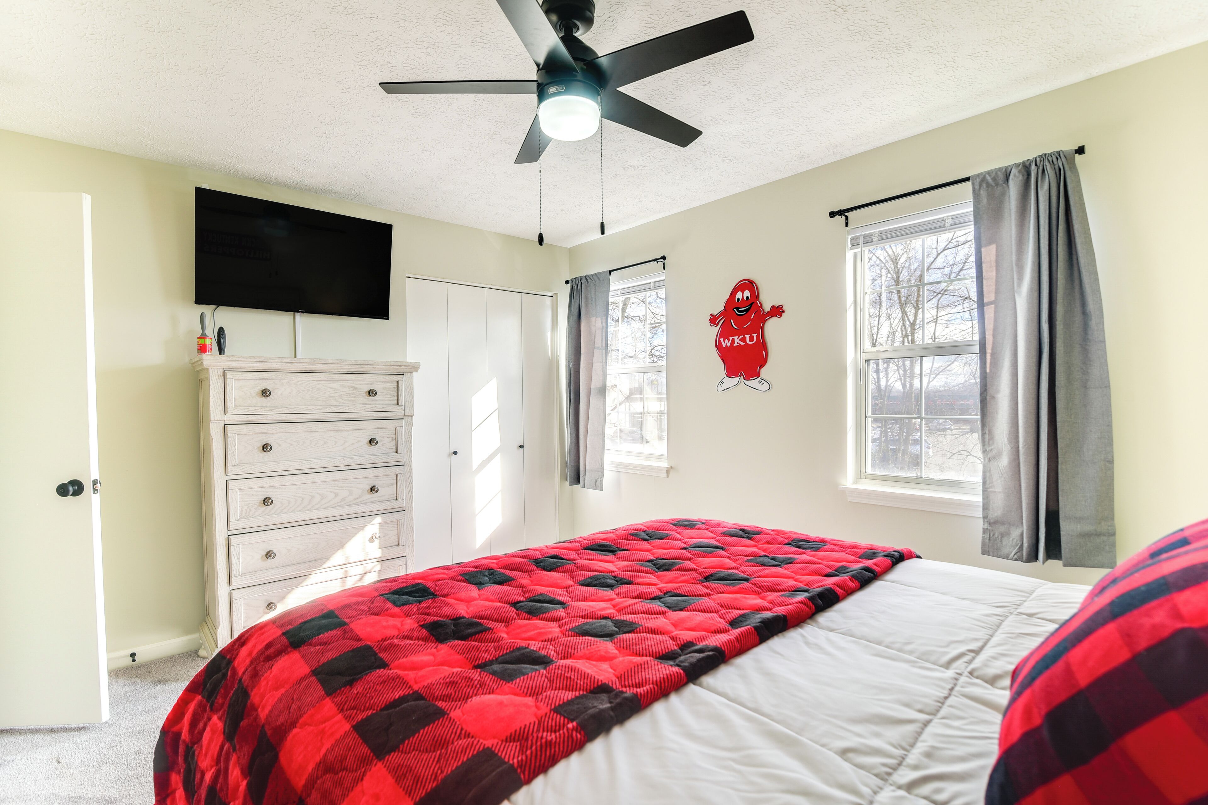 Ein Schlafzimmer mit Bett, Deckenventilator, Wand-Fernseher, Kommode, Fenster mit Vorhängen und einer roten Maskottchen-Dekoration.