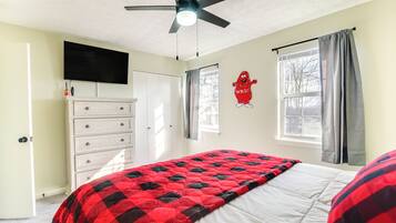 Ein Schlafzimmer mit Bett, Deckenventilator, Wand-Fernseher, Kommode, Fenster mit Vorhängen und einer roten Maskottchen-Dekoration.