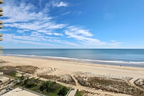 Blick auf einen Strand mit blauem Himmel, Sandstrand und einem eingezäunten Bereich mit Wegen.