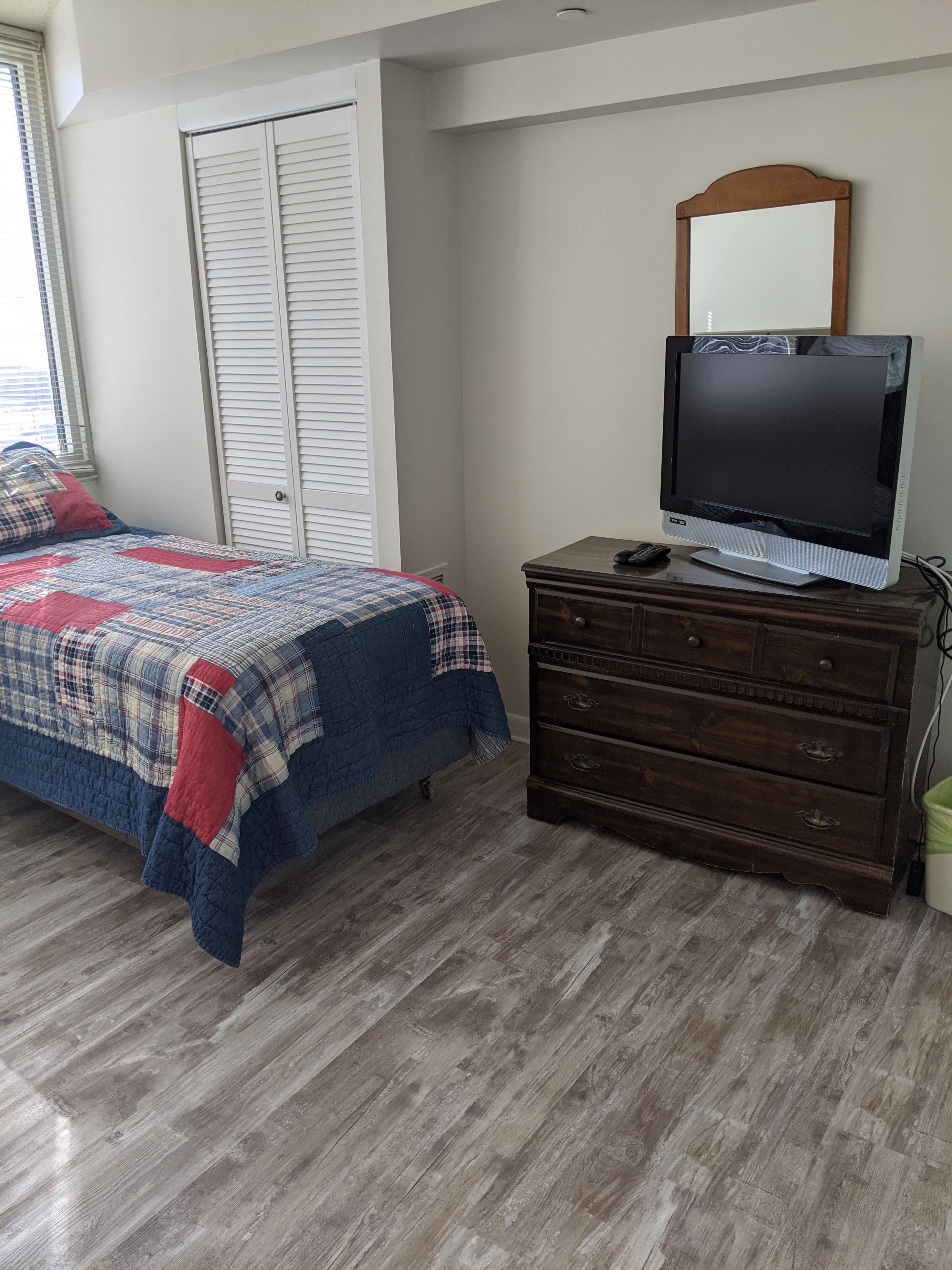 Une chambre à coucher comprenant un lit, une commode, un téléviseur et une fenêtre avec des stores.