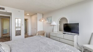 A bedroom with a bed, a TV, a dresser, a chair, and a door leading to a bathroom.