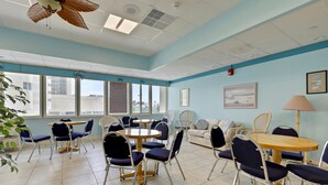 A room with multiple seating arrangements including tables and chairs, large windows, and a ceiling fan.
