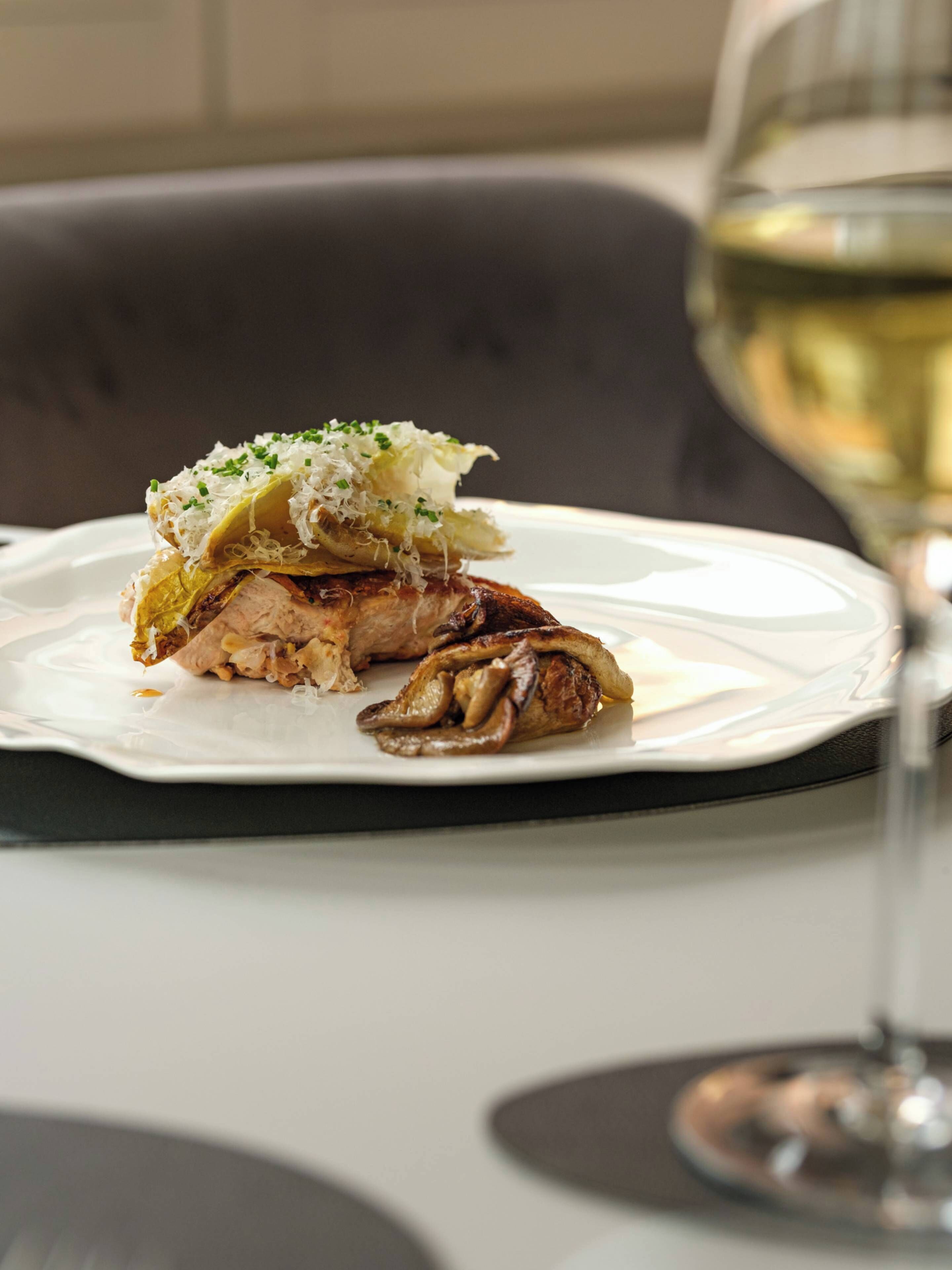 A plate with a piece of meat and mushrooms, garnished with shredded cheese and herbs, on a white plate.