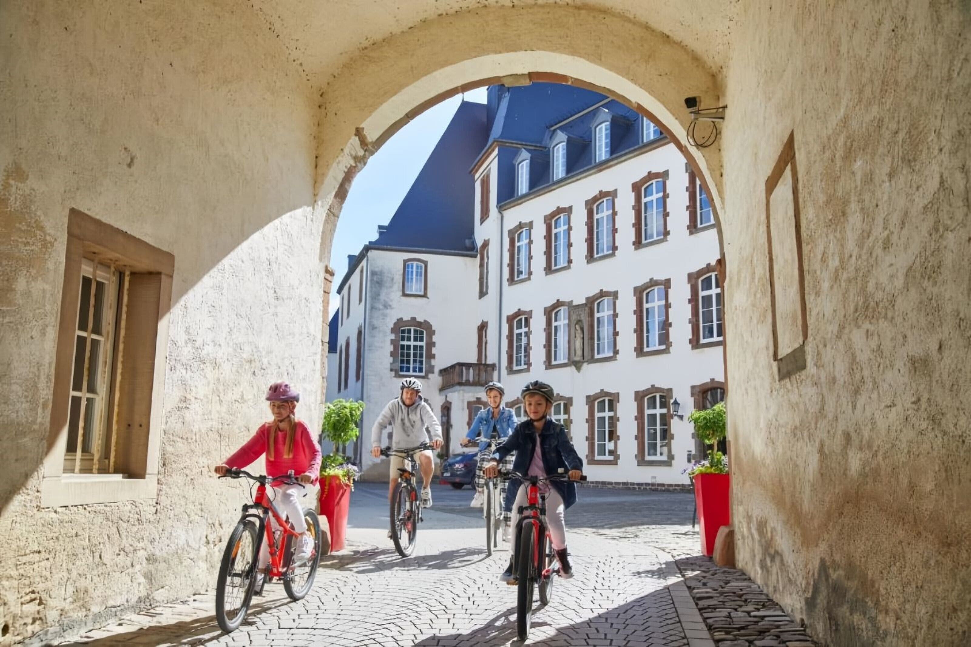 Un grupo de personas pedaleando a través de un arco de piedra con edificios al fondo.