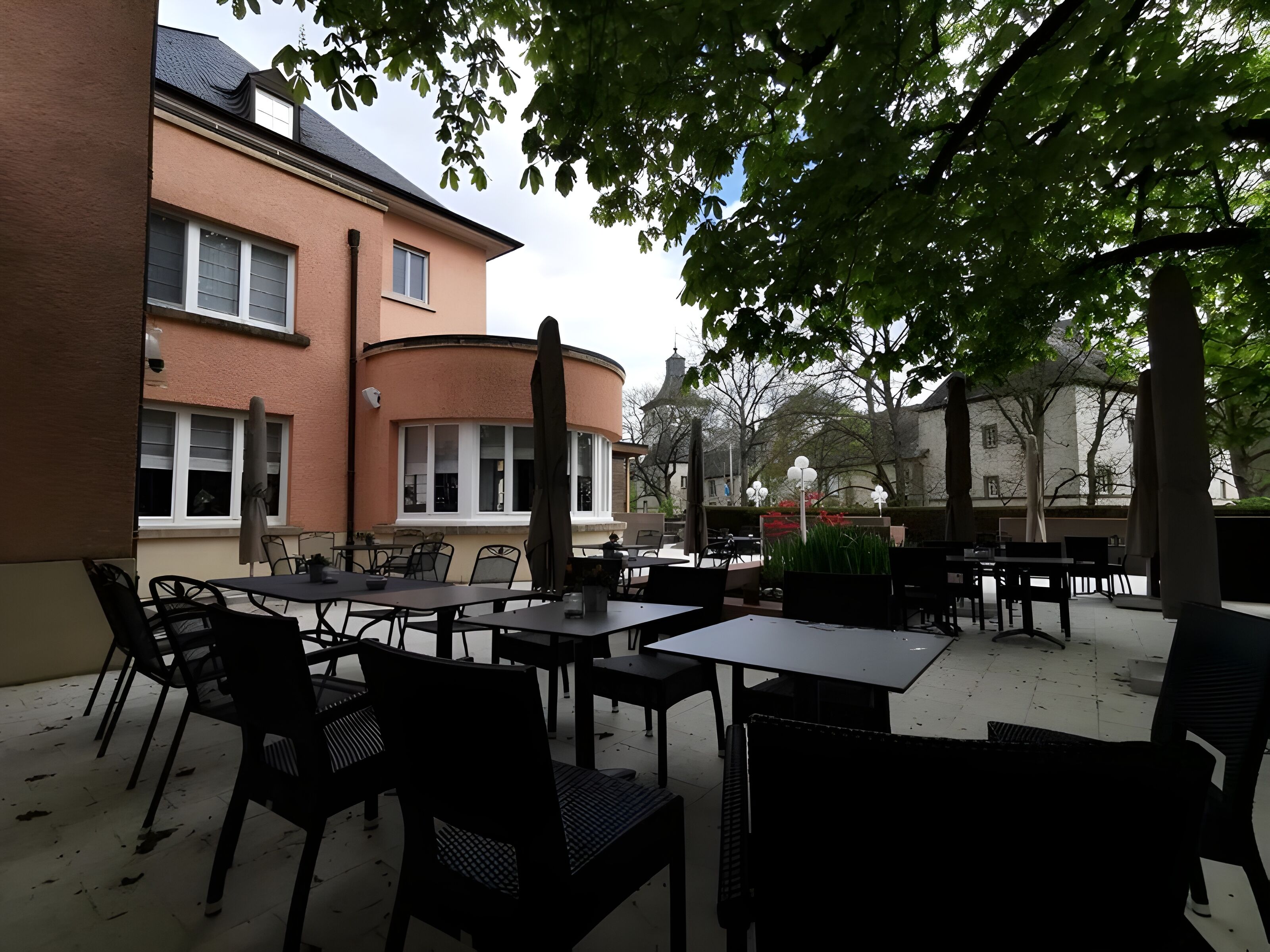 An outdoor seating area with tables and chairs, a building with a curved facade, and a tree providing shade.