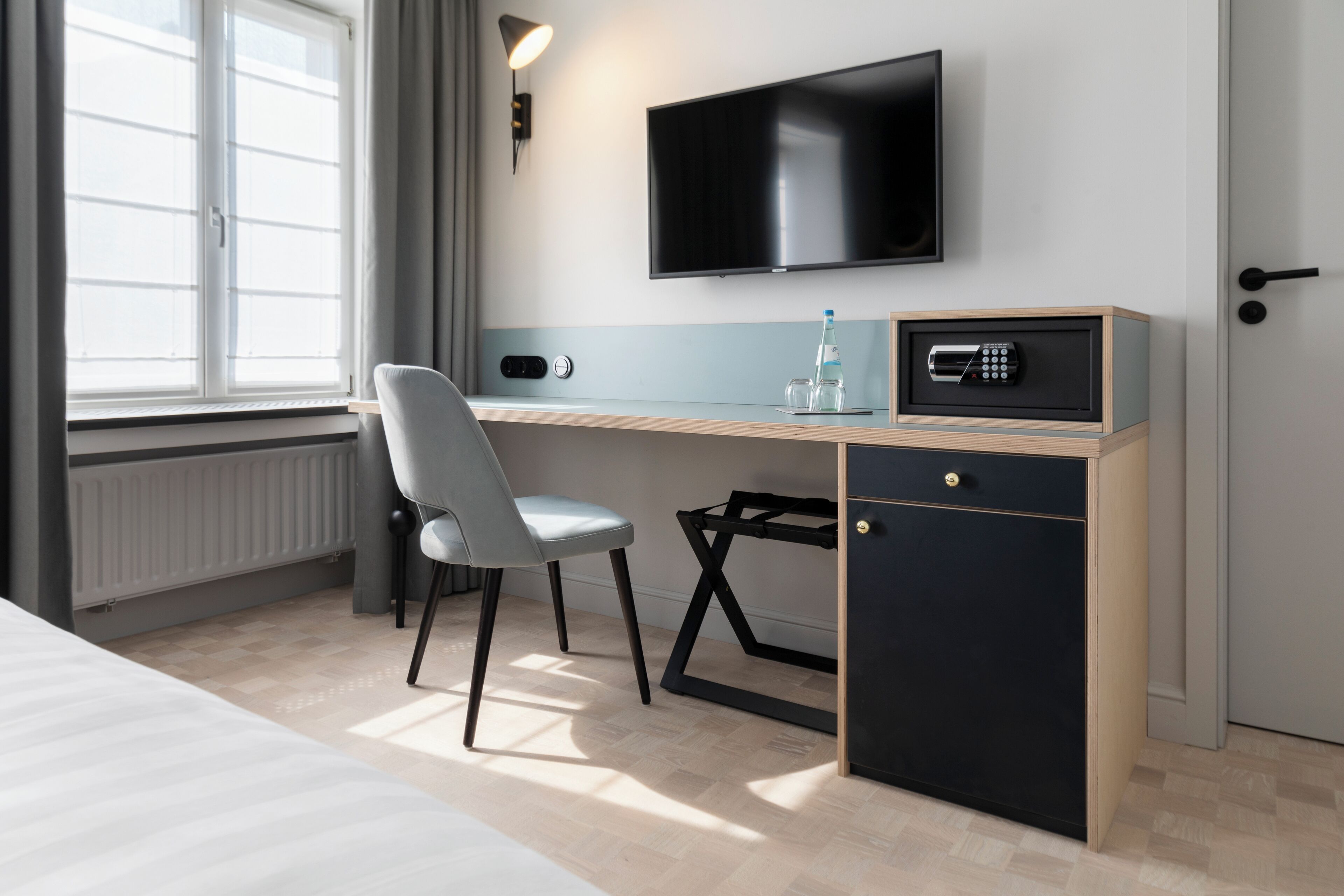 A hotel room with a bed, a desk with a chair, a TV mounted on the wall, a safe, and a window with curtains.