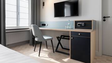 A hotel room with a bed, a desk with a chair, a TV mounted on the wall, a safe, and a window with curtains.