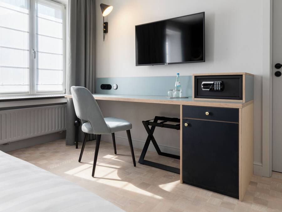 A hotel room with a bed, a desk with a chair, a TV mounted on the wall, a safe, and a window with curtains.